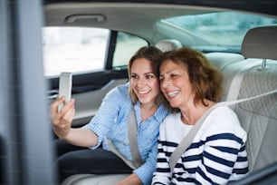 Adultos jóvenes y mayores alegres con teléfonos inteligentes sentados en los asientos traseros del automóvil, tomando selfies.