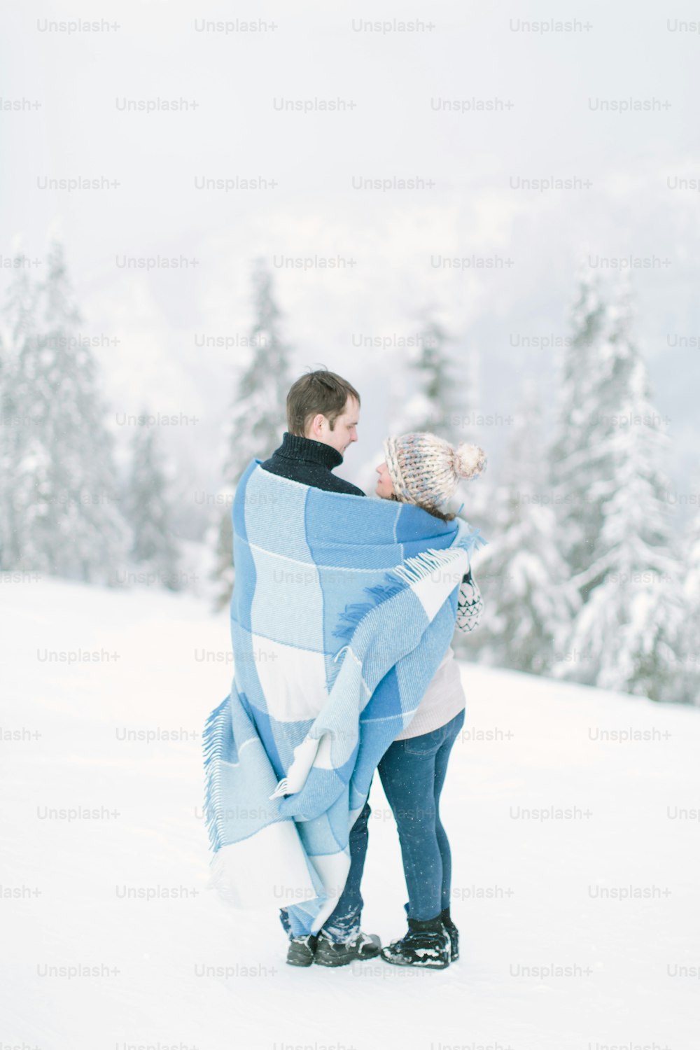 Young couple in love hugging and kissing in winter forest, covered with blue warm blanket..