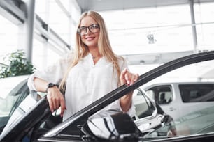 Cheerful customer. Young manager in the auto shop stands against perfectly polished black car.