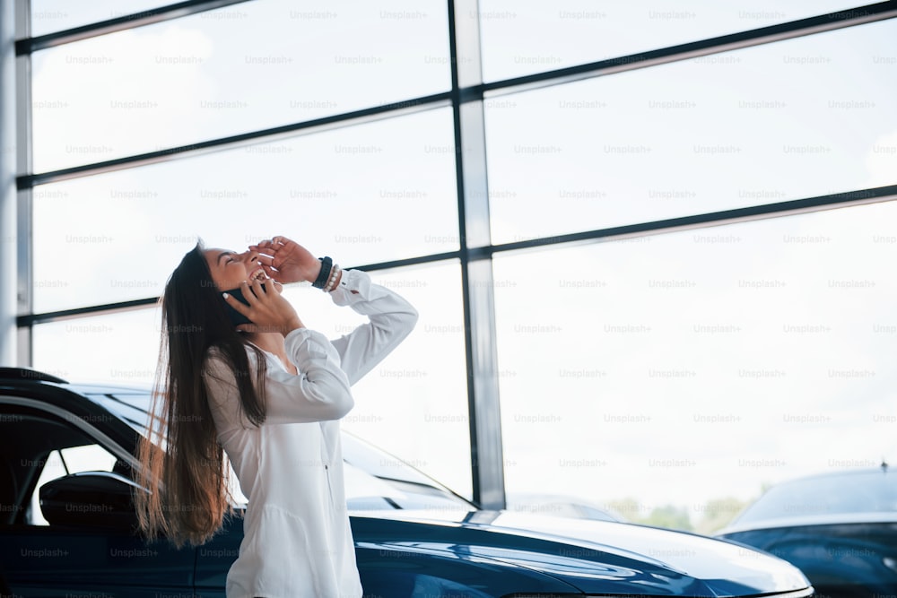 Having conversation by the phone. Young woman in white official clothes stands in front of blue automobile indoors.