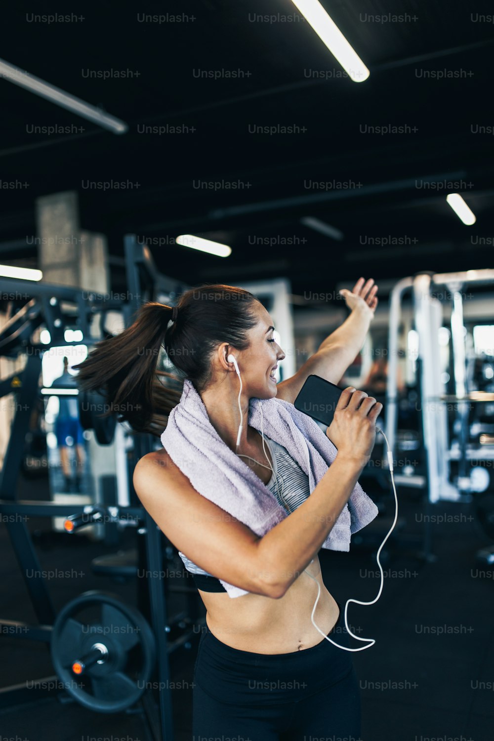 Young attractive woman after successful workout in modern fitness gym dancing, smiling and singing. She listening to music with wired earbuds and smart phone.