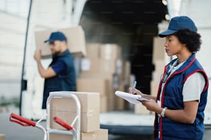 아프리카계 미국인 여성 노동자가 클립보드에 글을 쓰고 배달 목록을 살펴보는 동안 동료가 트럭에서 상자를 내리는 동안.