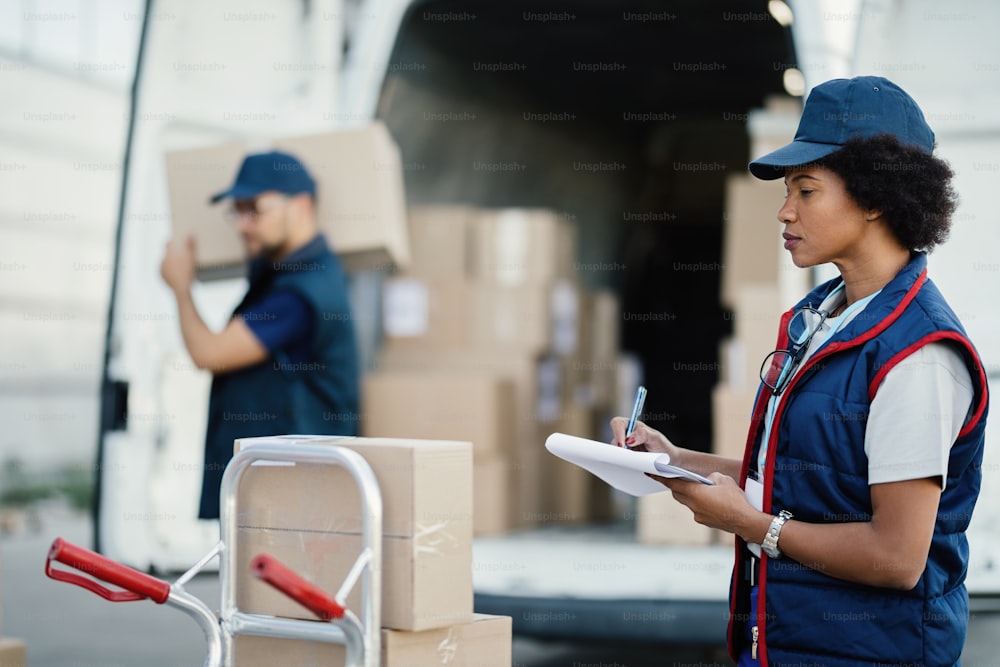 アフリカ系アメリカ人の女性労働者がクリップボードに書き込み、同僚がトラックから箱を降ろしている間に配達リストに目を通す。