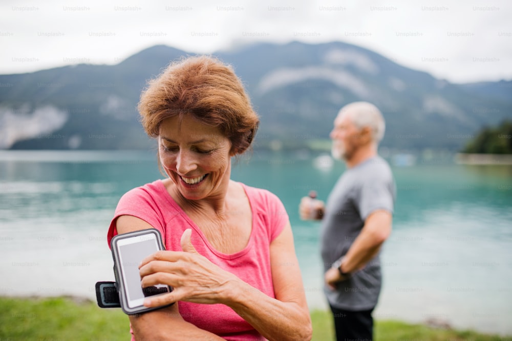 Senior pensioner couple runners with smartphone by lake in nature, resting.