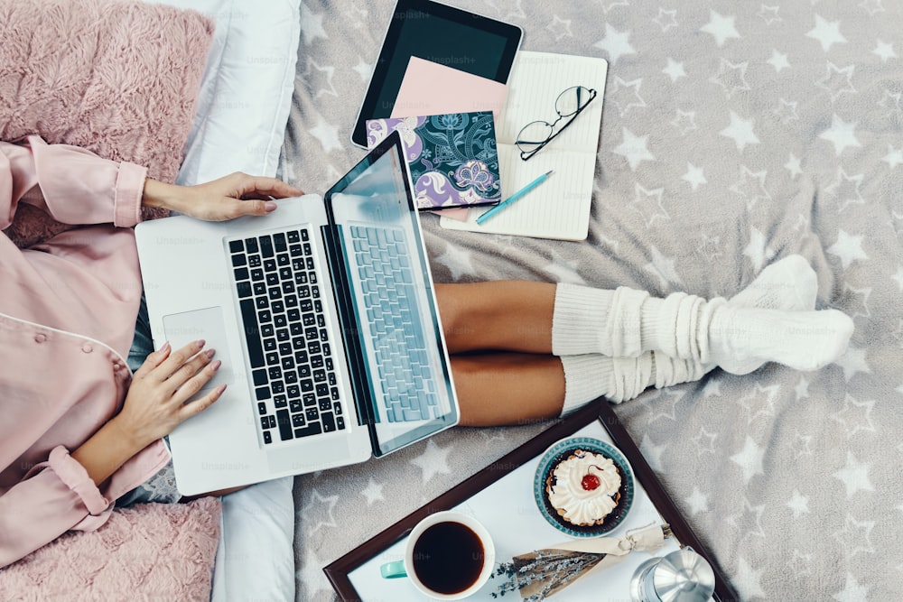 Close up top view of young woman in pajamas using laptop while resting in bed at home