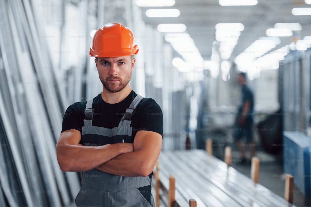 Stands with arms crossed. Portrait of male industrial worker indoors in factory. Young technician with orange hard hat.