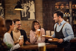 Groupe de jeunes gens heureux buvant de la bière pendant que le serveur apporte de la nourriture à leur table dans une taverne.