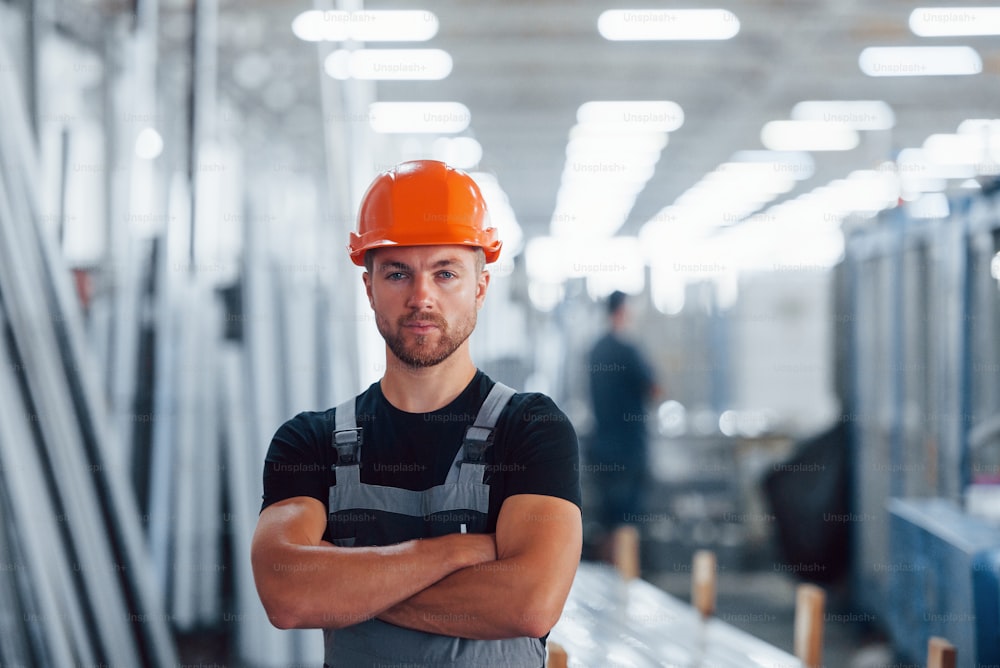 腕を組んで立つ。工場内の屋内での男性産業労働者の肖像画。オレンジ色のヘルメットをかぶった若い技術者。