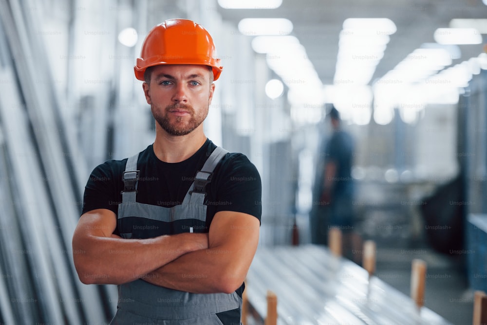 Stands with arms crossed. Portrait of male industrial worker indoors in factory. Young technician with orange hard hat.