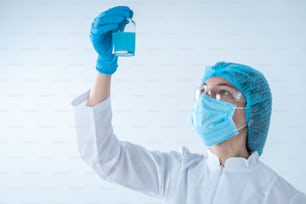Low angle view of scientist woman making clinical experiment, searching vaccine solution, holding medical glass bottle with blue liquid, checking sample, standing isolated on white background