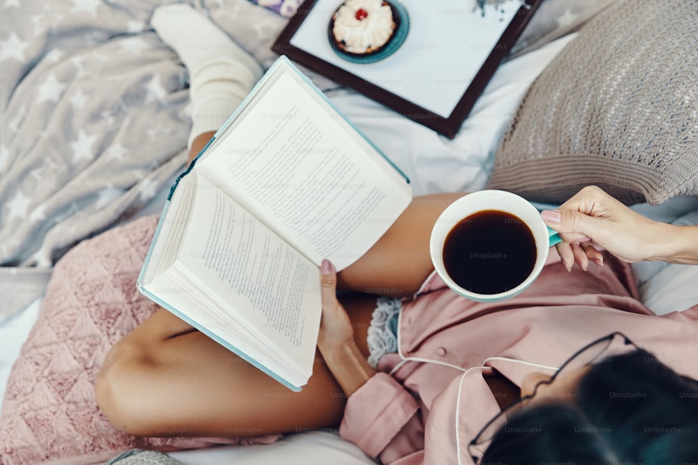Vista superior de una hermosa mujer joven en pijama leyendo un libro y disfrutando del café de la mañana mientras descansa en la cama en casa