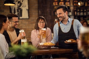 Pequeno grupo de amigos felizes bebendo cerveja enquanto o garçom está servindo-lhes um lanche em uma taverna.