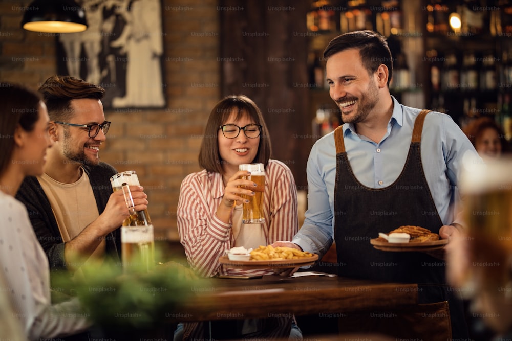 Kleine Gruppe glücklicher Freunde, die Bier trinken, während der Kellner ihnen einen Snack in einer Taverne serviert.
