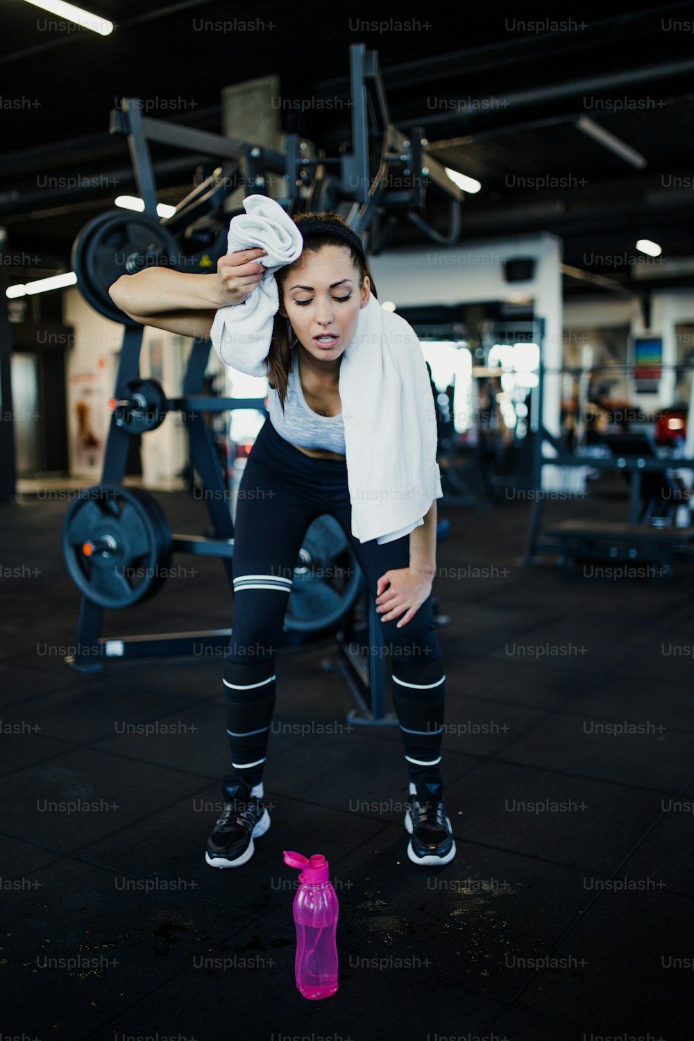 Young exhausted woman relaxing in fitness gym after successful workout wipes sweat from the forehead with white towel while holding water bottle in other arm.