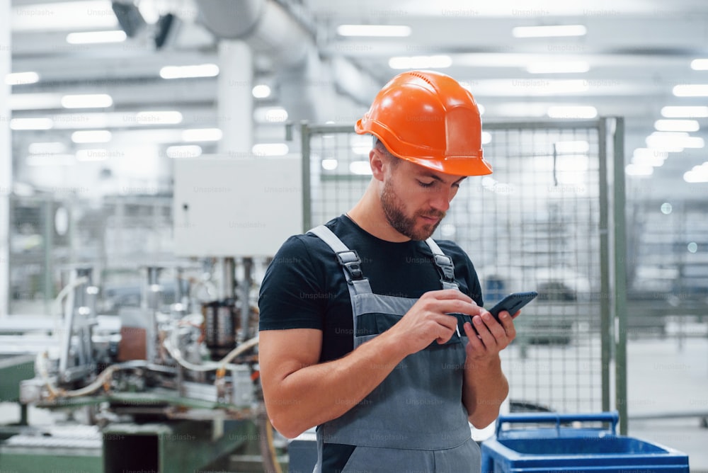 Smartphone in hands. Industrial worker indoors in factory. Young technician with orange hard hat.