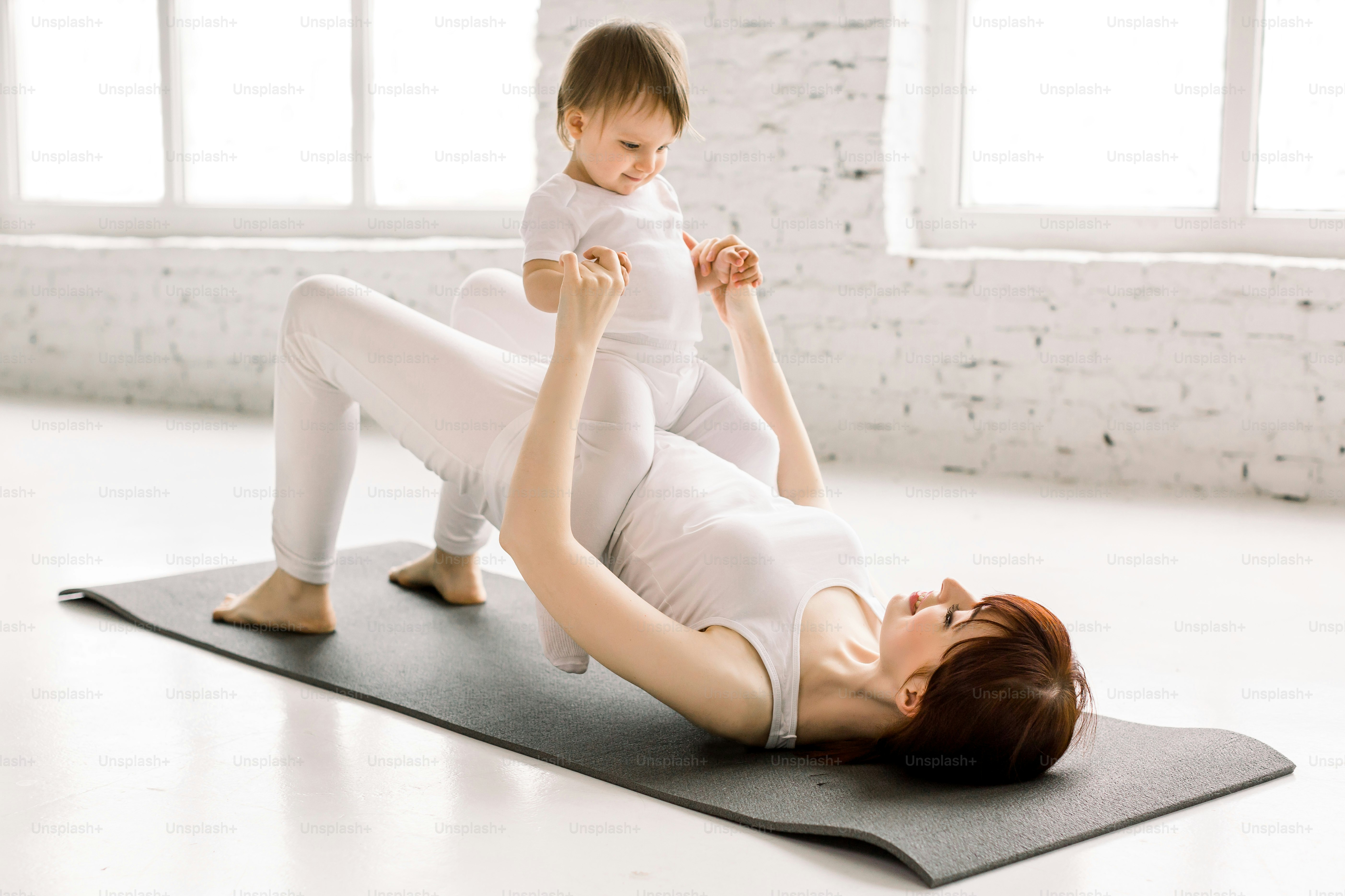 Young happy mother working out, doing butt bridge exercise, wearing white sportswear, little baby girl on her tummy, fitness, postnatal yoga. Healthy lifestyle concept.