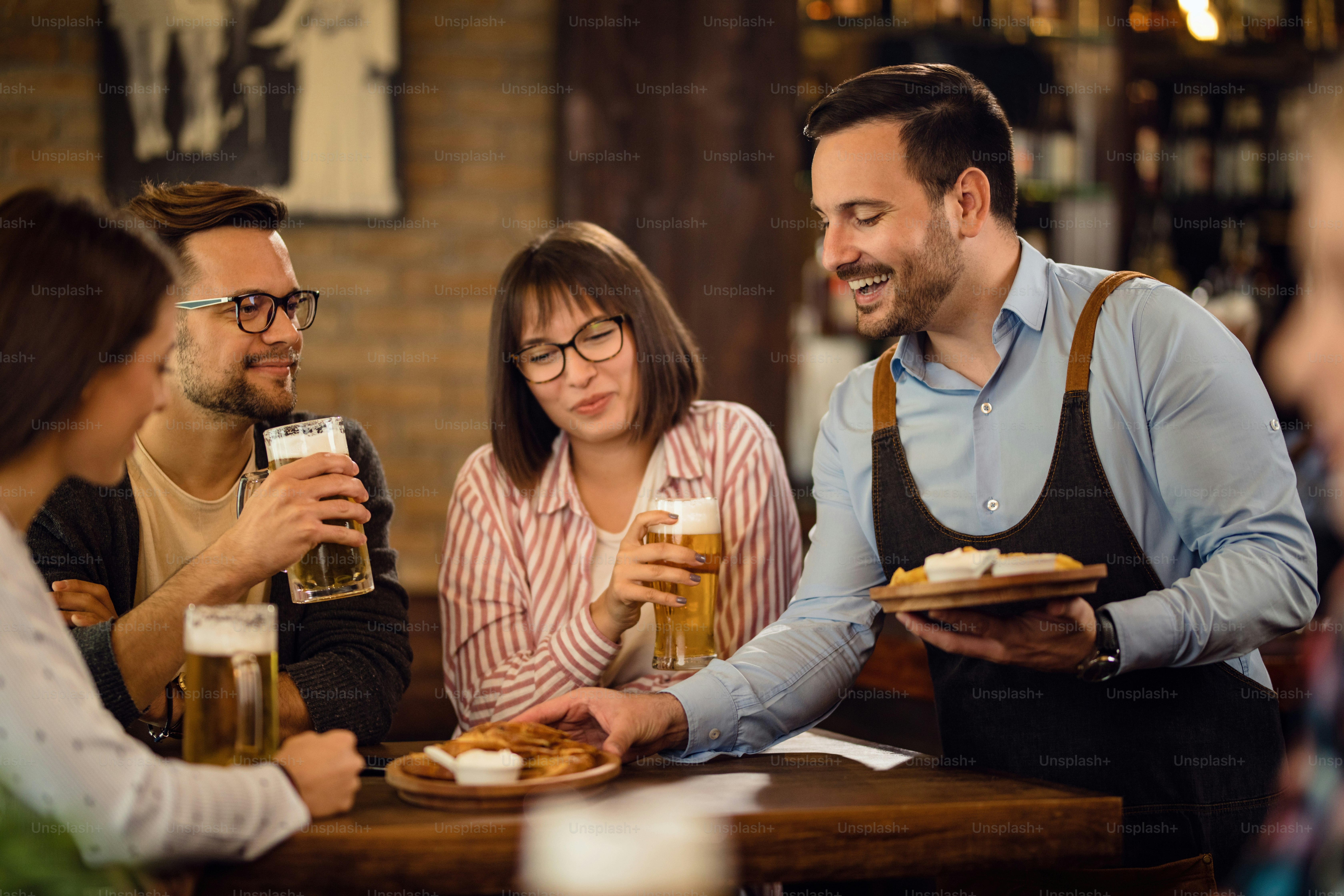 ウェイターがパブのテーブルで料理を提供している間、ビールを飲みながら楽しんでいる幸せな友人のグループ。