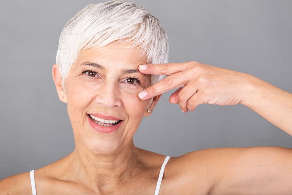 Portrait of smiling senior woman with perfect skin showing victory sign near eye on grey background. Closeup face of mature woman showing successful results after anti-aging wrinkle treatment.