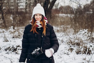 stylish hipster traveler girl with old photo camera exploring in snowy woods in winter. happy woman walking in cold forest. space for text. atmospheric moment. wanderlust
