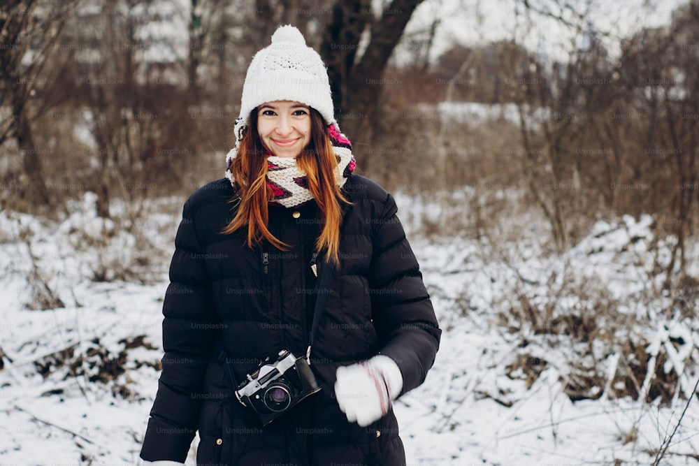 冬の雪に覆われた森を探索する古い写真カメラを持つスタイリッシュなヒップスターの旅行者の女の子。寒い森の中を歩く幸せな女性。テキスト用のスペース。雰囲気のある瞬間。放浪癖