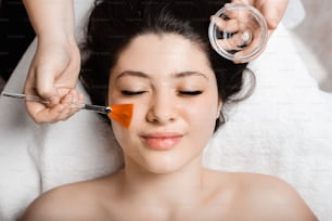 Upper view of a beautiful woman leaning with closed eyes while cosmetologist is applying a skin care mask on her face.