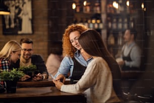 Young waitress assisting to a woman with choosing the order from a menu in a bar.