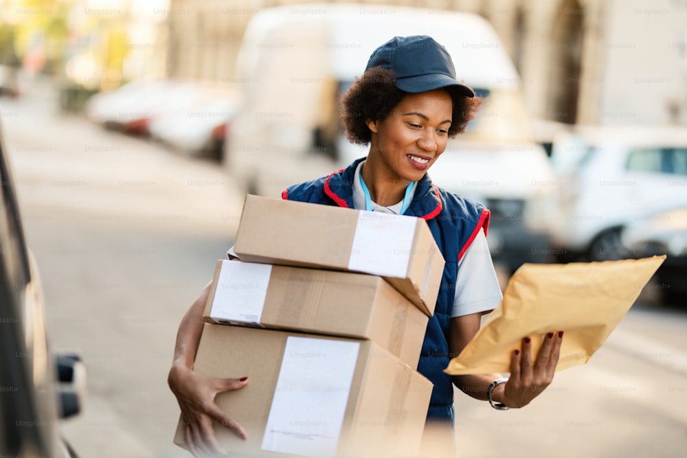 Repartidora afroamericana feliz leyendo la dirección en un paquete mientras hace la entrega en la ciudad.