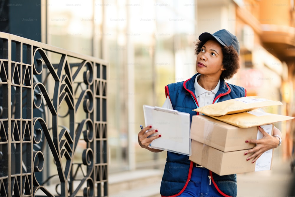Trabajadora postal afroamericana cargando paquetes mientras los entrega en un distrito residencial.