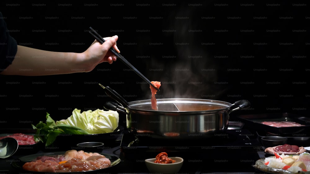 Plan recadré d’une femme mangeant du Shabu-Shabu dans une fondue avec de la viande fraîche tranchée, des fruits de mer et des légumes sur fond noir, cuisine japonaise