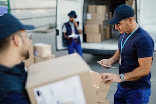 Young delivery man using touchpad and checking packages while organizing a  delivery with his coworkers.