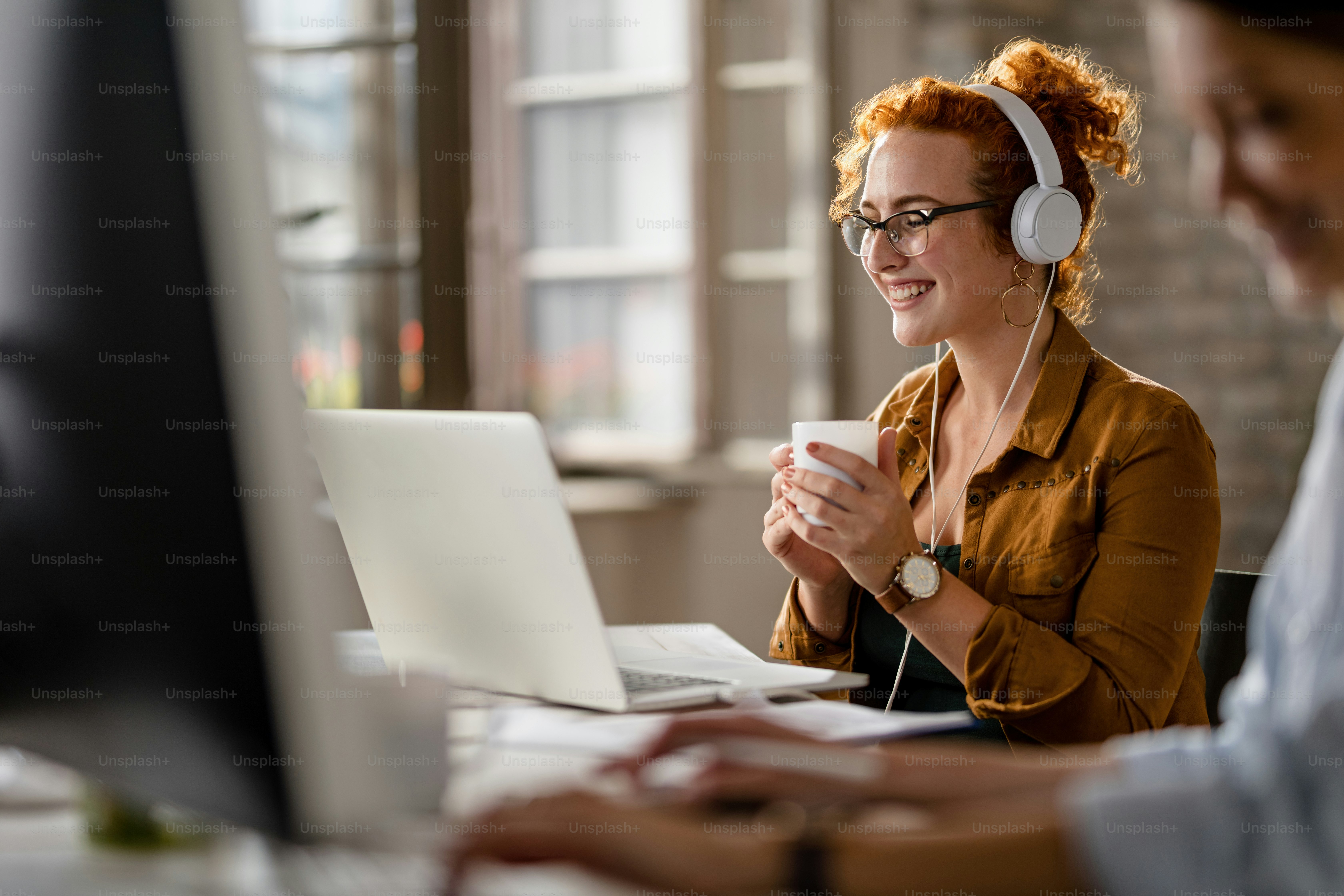 Feliz empresaria con auriculares mientras navega por la red en una computadora portátil y bebe café en la oficina.