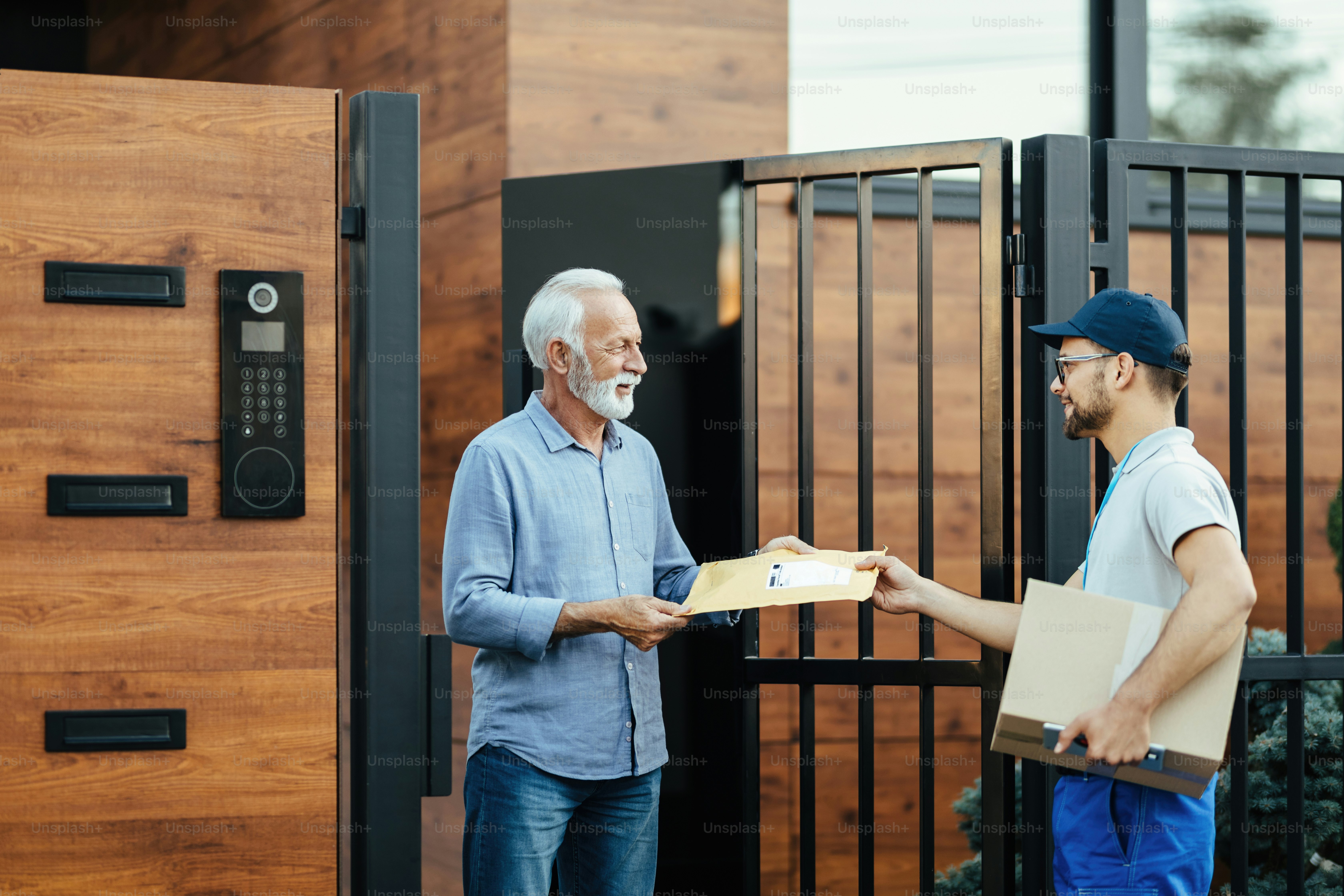Young postal worker making home delivery and giving a package to senior customer at front gate.