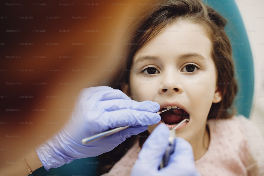 Gros plan d’une charmante petite fille assise dans un siège de stomatologie avec la bouche ouverte pendant que la stomatologie pédiatrique fait un examen des dents.