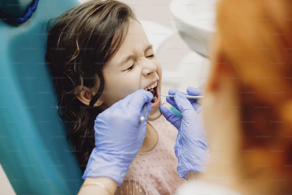 Charmante petite fille assise dans un siège de stomatologie avec les yeux fermés criant pendant que le médecin nettoie les dents des caries.