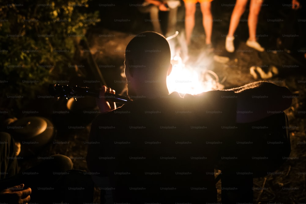 Hipster man playing on acoustic guitar and singing song at big bonfire at camp, chilling with friends in the forest. Group of people relaxing at fire in the evening, camping near lake