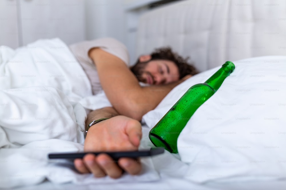 Texting while drunk concept. Young man lying in bed deadly drunken holding empty bottle of booze. intoxicated with alcohol . Alcoholism habitual drunkenness pernicious habit concept