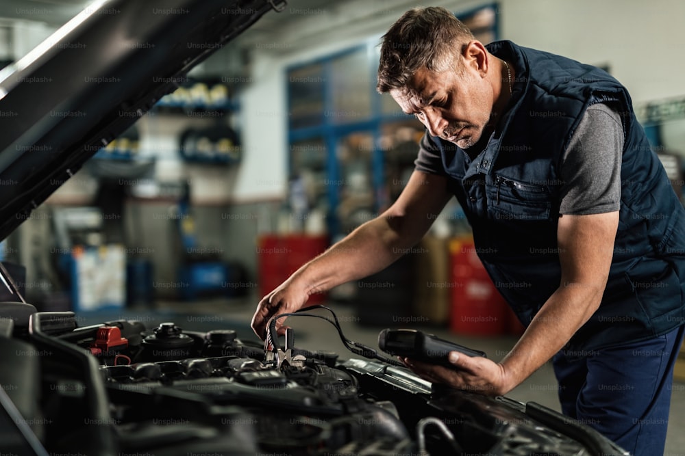 Auto mechanic using diagnostic tool while checking car battery voltage in a workshop.