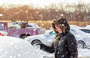Junge Frau, die an einem kalten Wintertag bei Schneefall den Schnee von seinem Auto abbürstet