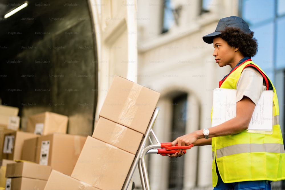 African American delivery woman with hand truck unloading packages from a truck.