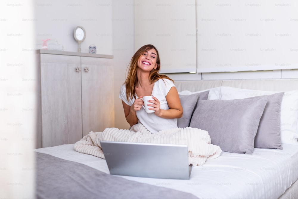 Mujer joven bebiendo café en casa en su cama y revisando su computadora portátil. Hermosa joven bebiendo café en casa en su cama mientras trabaja en su computadora portátil. Revisar el correo electrónico por la mañana