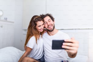 Young Couple In Bed Taking Selfie On Mobile Phone, Young happy beautiful couple on the bed making self portrait by smartphone and hugging,