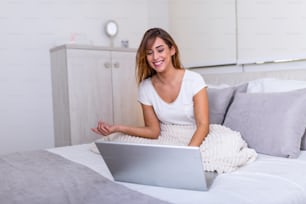 Portrait of a young smiling woman sitting with laptop on bed. Happy casual beautiful woman working on a laptop laying on the bed in the house. Freelancer woman working on her laptop at home