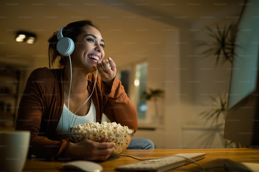 Low angle view of young woman having fun while eating popcorn and watching movie online on desktop PC at home.