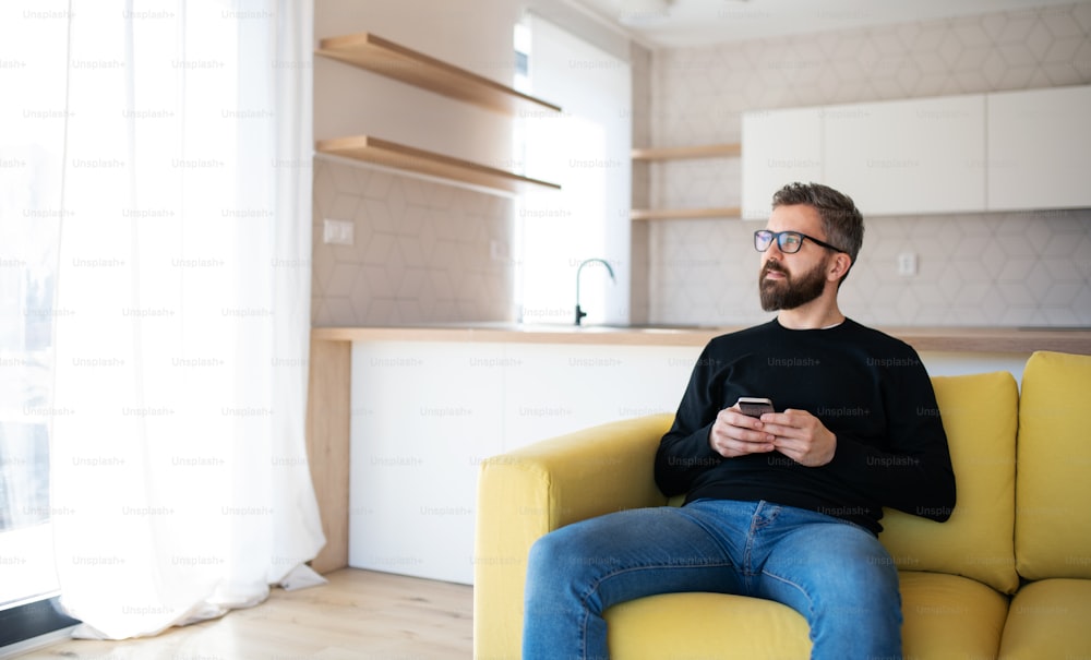 Un homme mûr heureux assis sur un canapé dans une maison non meublée, tenant un smartphone.
