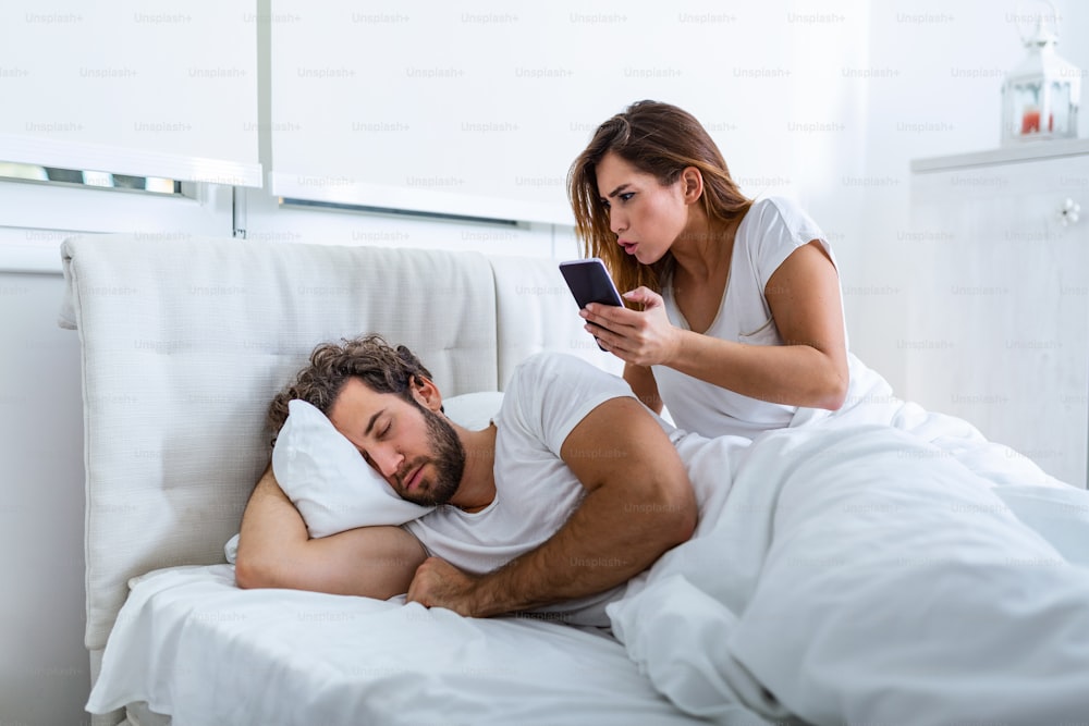 여자는 질투심이 많고 의심스럽고 침실에서 자고있는 동안 파트너의 스마트 폰을 염탐합니다. 아내는 남편이 자는 동안 남편의 전화를 염탐하고 있습니다. 불신, 질투의 개념