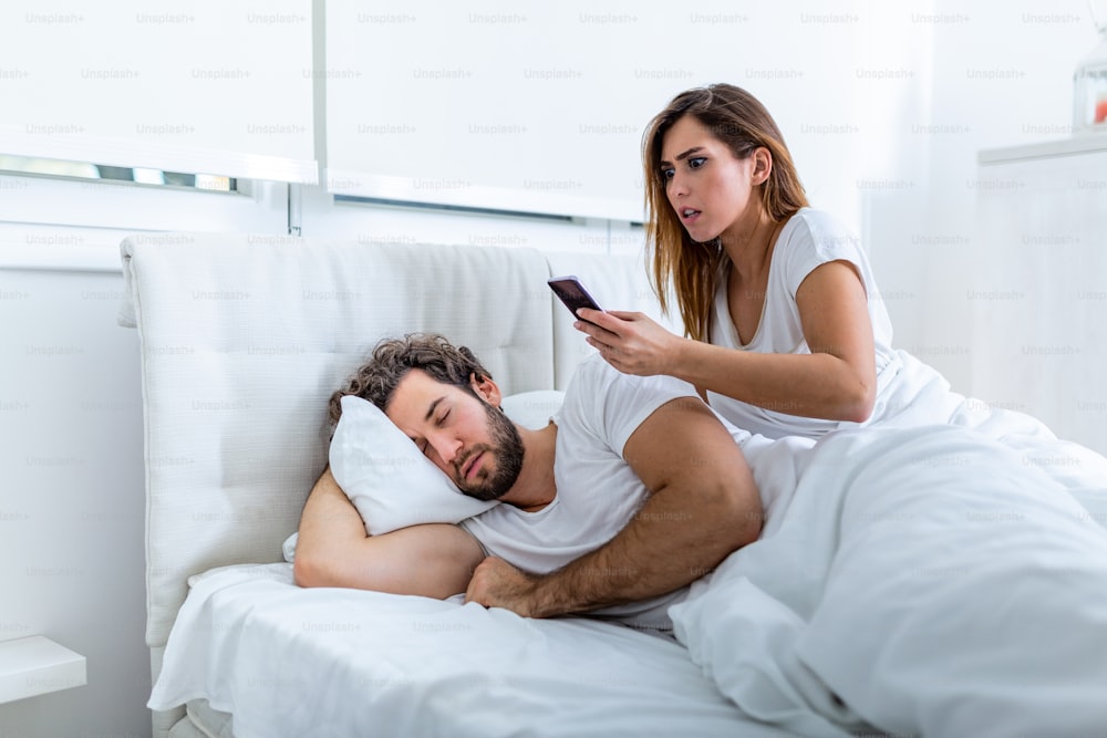 Jealous wife spying the phone of her partner while he is sleeping in a bed at home. Shocked jealous wife spying the phone of her husband while man sleeping in bed at home