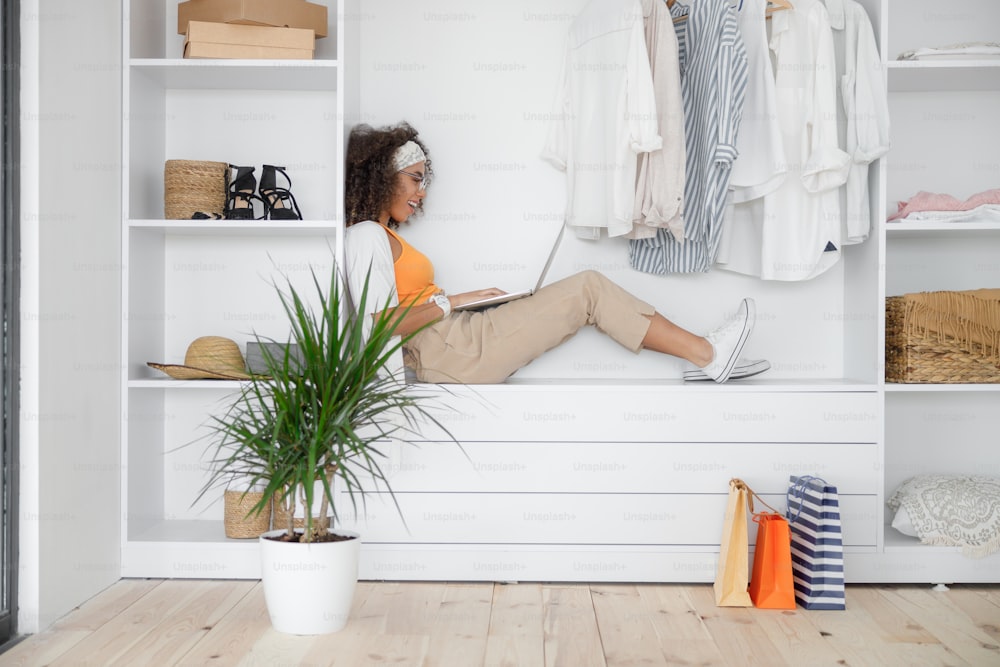 Smiling Afro American lady with laptop sitting in closet stock photo