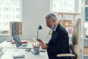Concentrated mature man in full suit using digital tablet while working in modern office