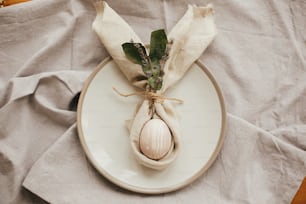 イースターテーブルの装飾、フラットレイ。イースターバニーナプキンに卵が入ったスタイリッシュなイースターブランチテーブルセッティング。ウサギの耳とナプキンにモダンな自然に染められたピンクの卵、皿にラベンダーの花