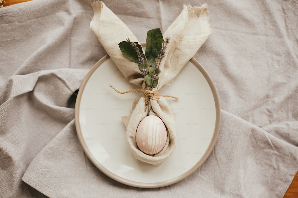 Easter table decorations, flat lay. Stylish Easter brunch table setting with egg in easter bunny napkin. Modern natural dyed pink egg on napkin with bunny ears, lavender flowers on plate